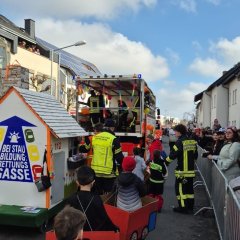 Kinder der Kinderfeuerwehr beim Umzug der Straßenfastnacht. Dahinter der Umzugswagen der Feuerwehr. Links und Rechts stehen am Straßenrand Zuschauer.