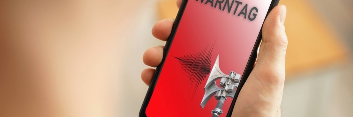 Eine Hand hält ein Smartphone. Auf dem roten Display steht das Wort Warntag. Der Hintergrund ist verschwommen.