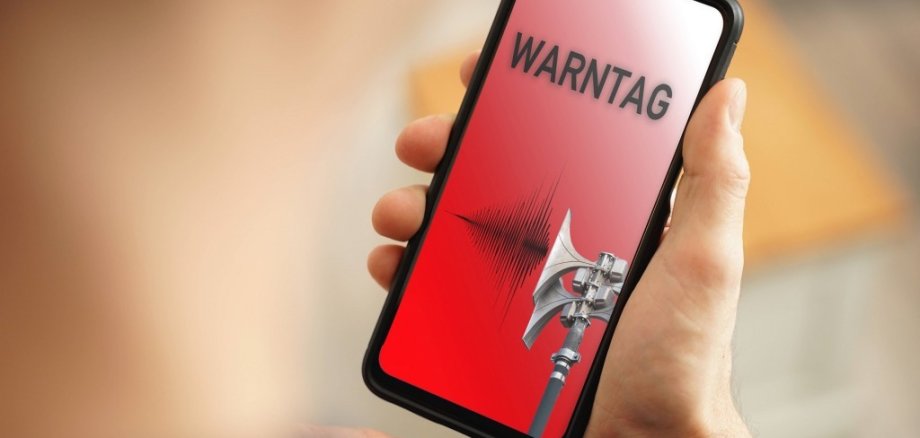 Eine Hand hält ein Smartphone. Auf dem roten Display steht das Wort Warntag. Der Hintergrund ist verschwommen.