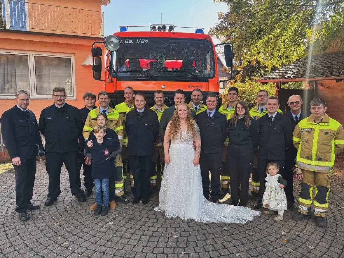 Gruppenbild einer Hochzeit. In der Mitte das Brautpaar. Darum Personen in Feuerwehruniformen. Dahinter ein Feuerwehrfahrzeug.