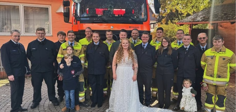 Gruppenbild einer Hochzeit. In der Mitte das Brautpaar. Darum Personen in Feuerwehruniformen. Dahinter ein Feuerwehrfahrzeug.