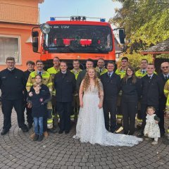 Gruppenbild einer Hochzeit. In der Mitte das Brautpaar. Darum Personen in Feuerwehruniformen. Dahinter ein Feuerwehrfahrzeug.