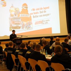 Vortrag im Saal der Feuerwehr-Jahreshauptversammlung 2025: Eine Person am Rednerpult spricht vor sitzendem Publikum, im Hintergrund eine Präsentation mit Feuerwehr-Slogan.
