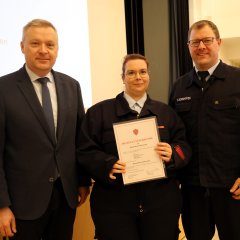 Drei Personen in feuerwehr- oder formeller Kleidung stehen nebeneinander; die mittlere Person hält eine Urkunde zur Feuerwehr-Ehrung.