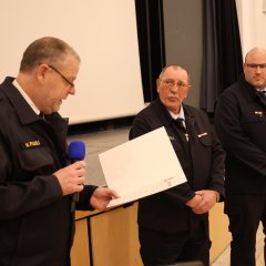 Drei Feuerwehrangehörige in Uniform stehen nebeneinander; der links stehende spricht ins Mikrofon und überreicht eine Urkunde.