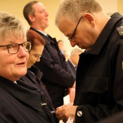 Zwei Feuerwehrleute in Uniform stehen nah beieinander; einer steckt dem anderen eine Auszeichnung an die Kleidung.