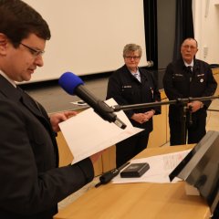 Bei der Jahreshauptversammlung der Feuerwehr Gernsheim 2025 liest ein Sprecher am Rednerpult vor, während zwei Uniformierte neben ihm stehen und zuhören.