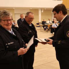 Bei der Feuerwehr‑Jahreshauptversammlung 2025 überreicht ein Feuerwehrangehöriger in Uniform einem anderen eine Urkunde, beide lächeln, im Saal sitzen weitere Gäste im Hintergrund.