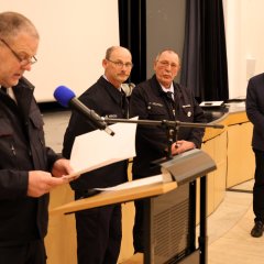 Bei der Jahreshauptversammlung der Feuerwehr Gernsheim 2025 liest ein Feuerwehrmann am Rednerpult vor, während zwei Feuerwehrleute und der Bürgermeister neben ihm stehen und zuhören.