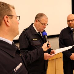 Bei der Jahreshauptversammlung der Feuerwehr Gernsheim 2025 liest ein Sprecher am Rednerpult vor, links und rechts stehen ein Mitglied der Feuerwehr und hören zu.