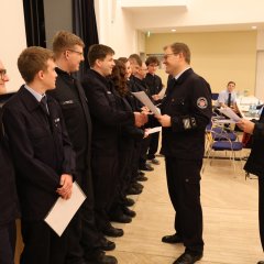 Zwei Feuerwehrleute stehen rechts und verteilen an neun in Reihe stehende Feuerwehrleute auf der linken Seite Urkunden.