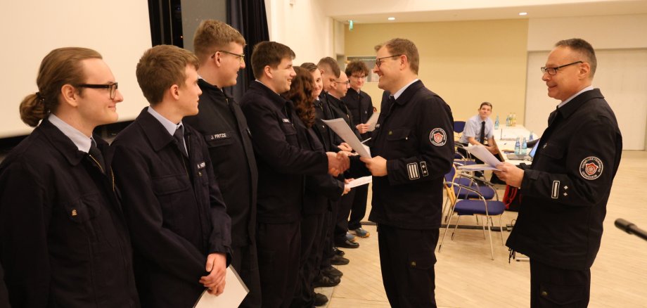 Zwei Feuerwehrleute stehen rechts und verteilen an neun in Reihe stehende Feuerwehrleute auf der linken Seite Urkunden.