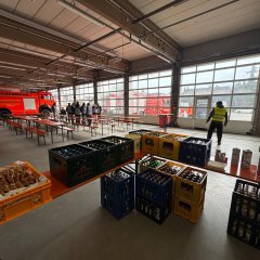 Fahrzeughalle der Feuerwehr. Die Fahrzeuge stehen vor dem Tor. Es sind viele Biergartengarnituren aufgestellt. Im Vordergrund eine Theke mit diversen Getränkekisten.
