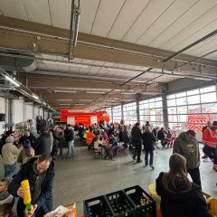 Fahrzeughalle der Feuerwehr. Die Fahrzeuge stehen vor dem Tor. Es sind viele Biergartengarnituren aufgestellt an denen Menschen sitzen. In der ganzen Halle stehen eine Vielzahl von Menschen. Im Vordergrund eine Theke mit diversen Getränkekisten.
