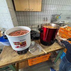 Zwei große Glühweinkocher mit Würstchen und eine Kiste mit Brötchen. Eine Frau schneidet gerade ein Brötchen auf.