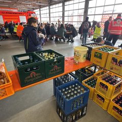 Fahrzeughalle der Feuerwehr. Es sind viele Biergartengarnituren aufgestellt an denen Menschen sitzen. In der ganzen Halle stehen eine Vielzahl von Menschen. Im Vordergrund eine Theke mit diversen Getränkekisten.