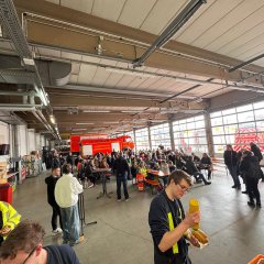Fahrzeughalle der Feuerwehr. Die Fahrzeuge stehen vor dem Tor. Es sind viele Biergartengarnituren aufgestellt an denen Menschen sitzen. In der ganzen Halle stehen eine Vielzahl von Menschen.