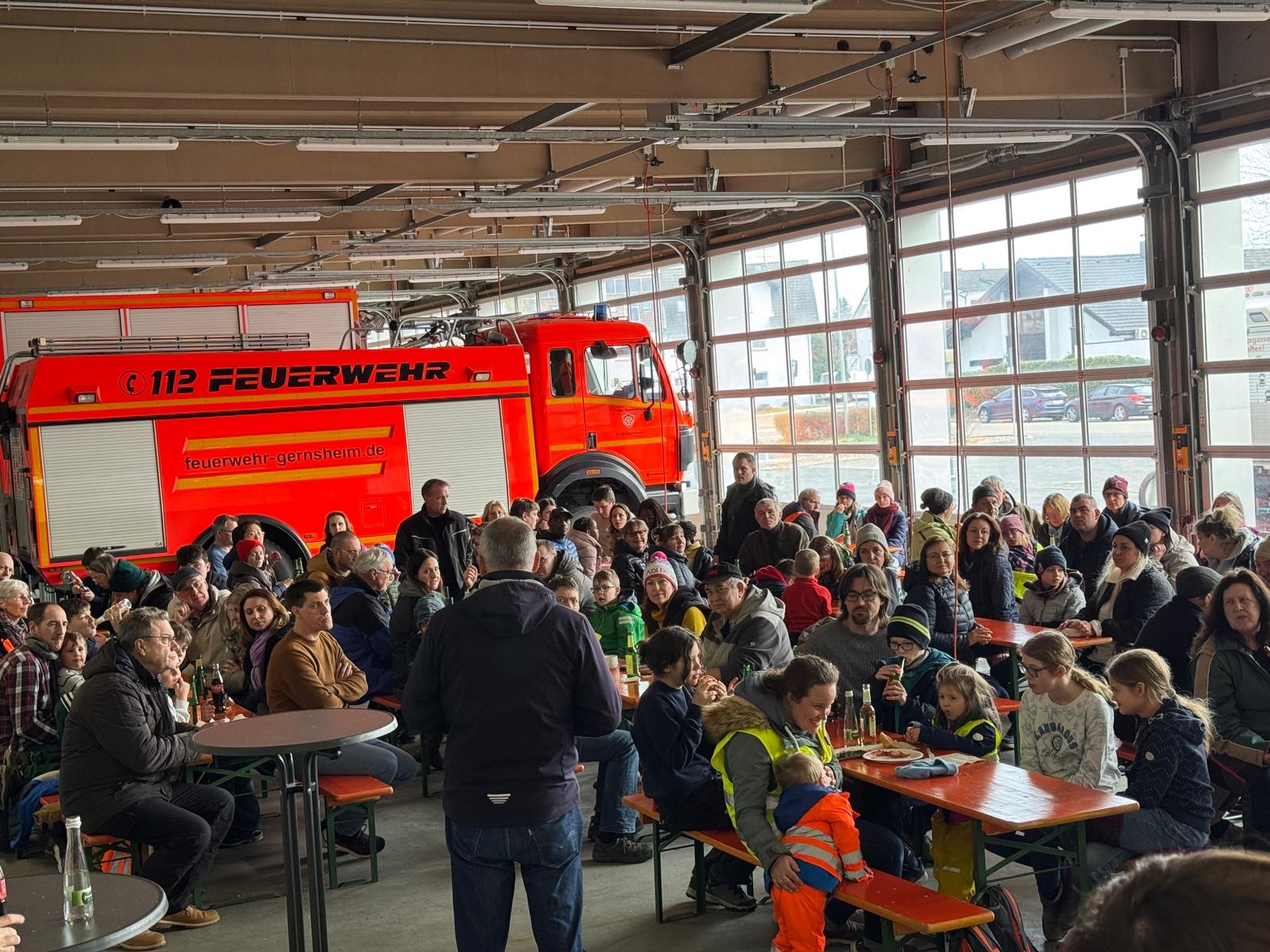 Fahrzeughalle der Feuerwehr. Die Fahrzeuge stehen vor dem Tor. Es sind viele Biergartengarnituren aufgestellt an denen Menschen sitzen. In der Mitte steht der Bürgermeister und hält eine Rede.