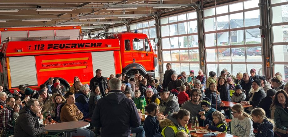 Fahrzeughalle der Feuerwehr. Die Fahrzeuge stehen vor dem Tor. Es sind viele Biergartengarnituren aufgestellt an denen Menschen sitzen. In der Mitte steht der Bürgermeister und hält eine Rede.