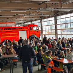 Fahrzeughalle der Feuerwehr. Die Fahrzeuge stehen vor dem Tor. Es sind viele Biergartengarnituren aufgestellt an denen Menschen sitzen. In der Mitte steht der Bürgermeister und hält eine Rede.