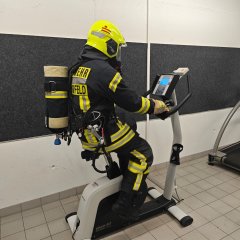 Feuerwehrangehöriger in Schutzkleidung und Atemschutzmaske auf einem Trimmrad in einer Übungshalle.