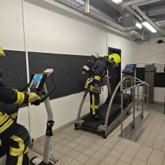Feuerwehrangehöriger in Schutzkleidung und Atemschutzmaske auf einem Laufband in einer Übungshalle.