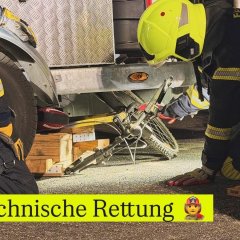 ZweiFeuerwehrleute in Einsatzkleidung knien an einem Anhänger während einer technischen Rettungsübung. Unter dem Anhänger befindet sich ein Fahrrad.