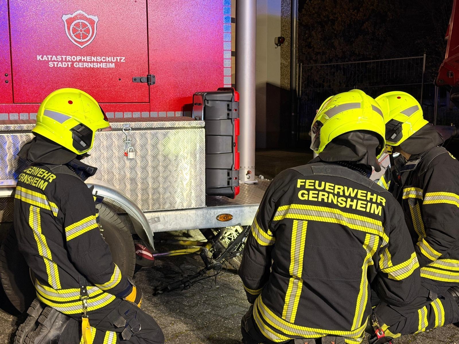 Drei Feuerwehrleute in Einsatzkleidung knien an einem Anhänger während einer technischen Rettungsübung. Unter dem Anhänger befindet sich ein Fahrrad.