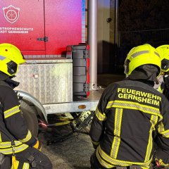 Drei Feuerwehrleute in Einsatzkleidung knien an einem Anhänger während einer technischen Rettungsübung. Unter dem Anhänger befindet sich ein Fahrrad.