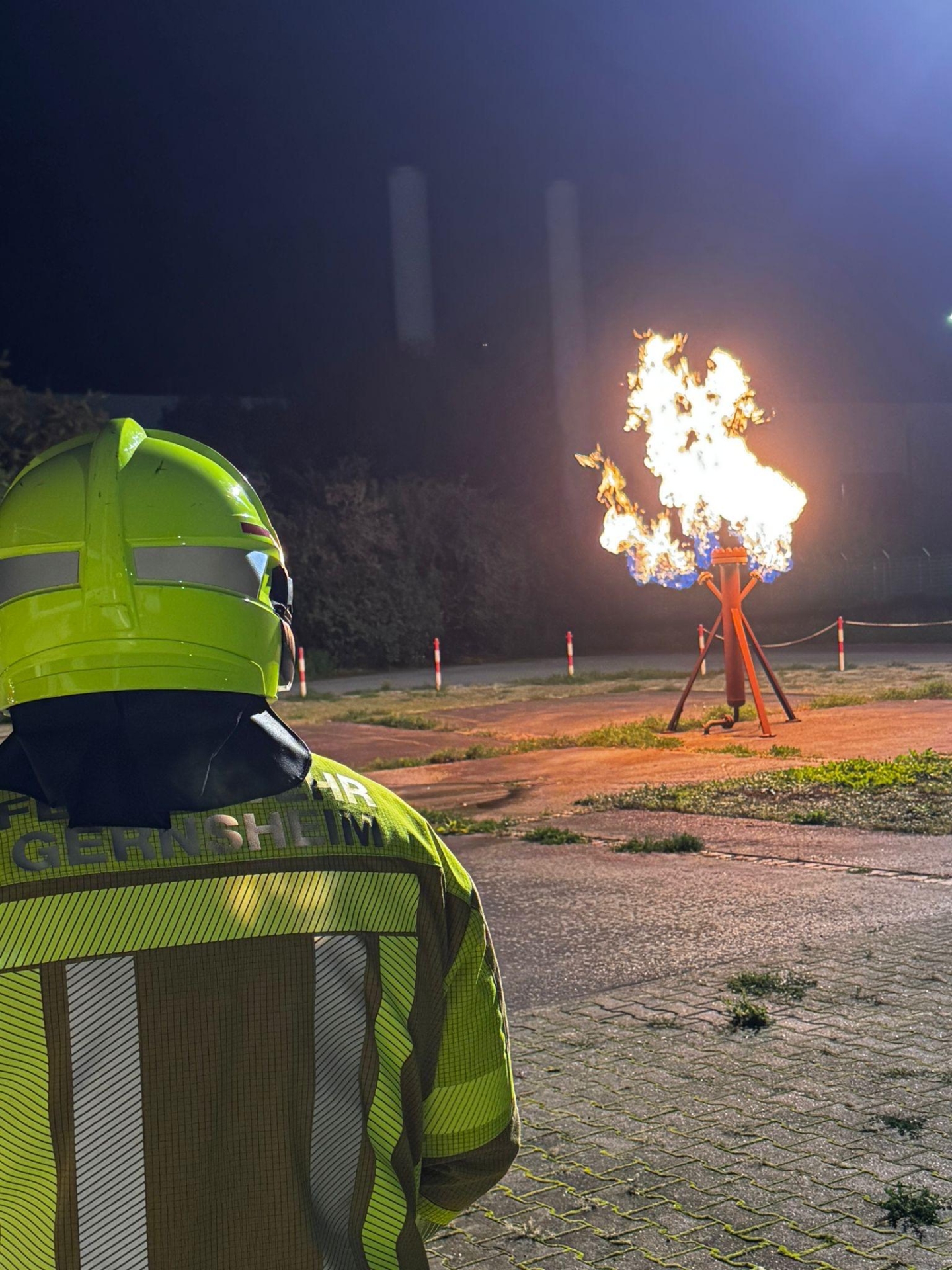 Von hinten der Kopf und Oberkörper eines Feuerwehrmannes. Im Hintergrund eine brennende Gassäule bei einer Übung. Es ist Nacht.