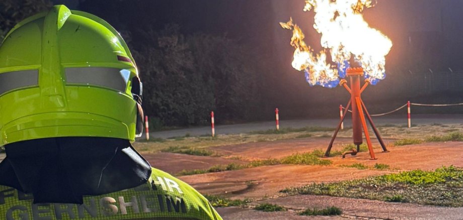 Von hinten der Kopf und Oberkörper eines Feuerwehrmannes. Im Hintergrund eine brennende Gassäule bei einer Übung. Es ist Nacht.