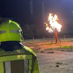 Von hinten der Kopf und Oberkörper eines Feuerwehrmannes. Im Hintergrund eine brennende Gassäule bei einer Übung. Es ist Nacht.