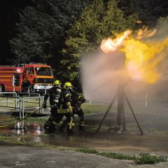 Drei Feuerwehrleute löschen eine brennende Gassäule bei einer Übung. Im Hintergrund ein Feuerwehrfahrzeug. Es ist Nacht. 