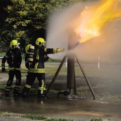 Drei Feuerwehrleute löschen eine brennende Gassäule bei einer Übung. Es ist Nacht.