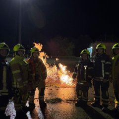Sechs Feuerwehrleute stehen in Reihe. In der Mitte dahinter ein Stück brennendes Gras nach einer Übung. Es ist Nacht.