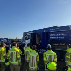 Feuerwehrleute in Schutzkleidung stehen vor einem blauen LKW. Ein Mann in blauer Arbeitskleidung spricht zu ihnen.
