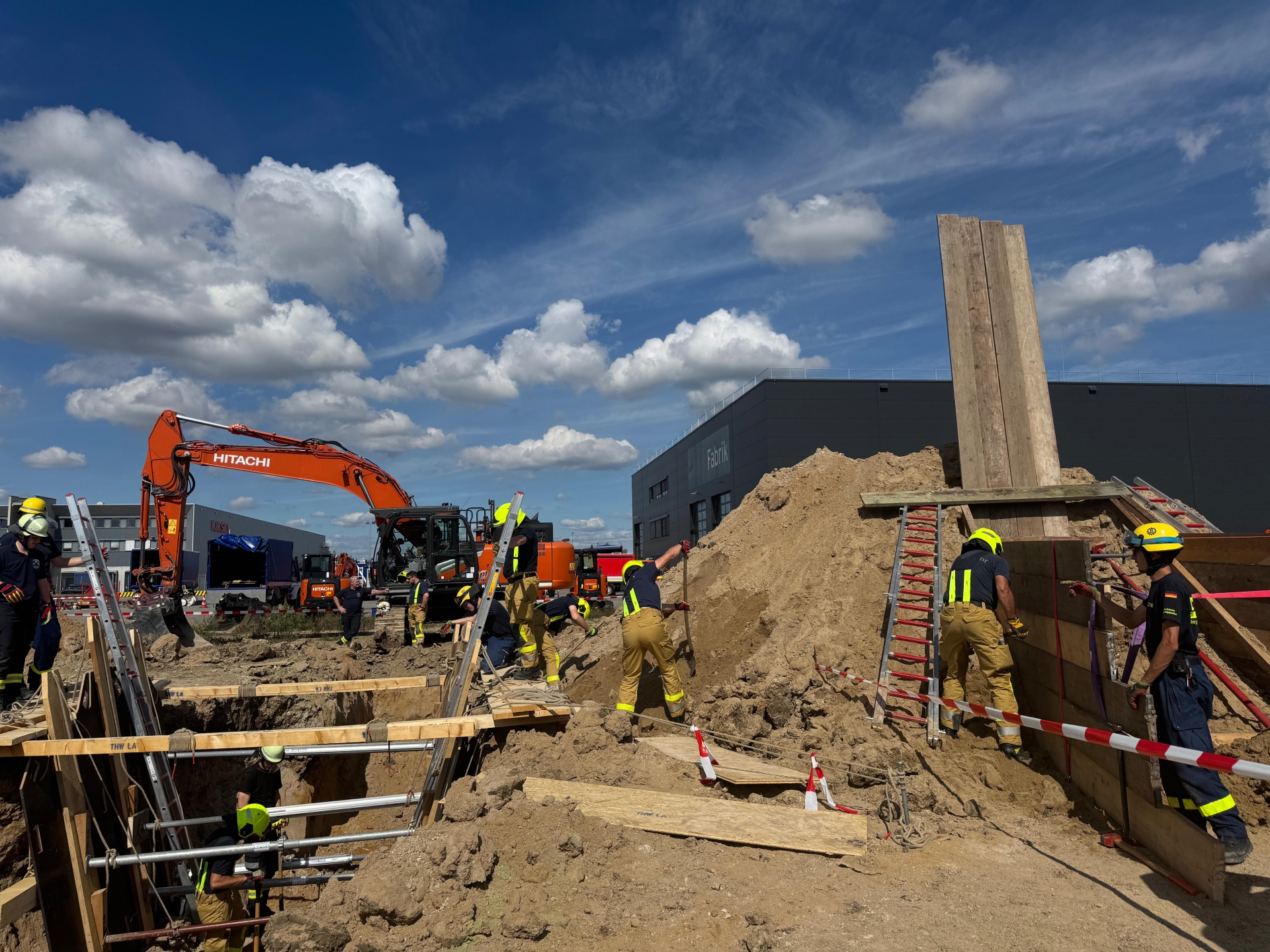 Feuerwehrleute in Schutzkleidung arbeiten gemeinsam an einer Übung im Bereich Technische Hilfeleistung am Bau. Im Hintergrund ein oragner Bagger.