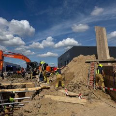 Feuerwehrleute in Schutzkleidung arbeiten gemeinsam an einer Übung im Bereich Technische Hilfeleistung am Bau. Im Hintergrund ein oragner Bagger.