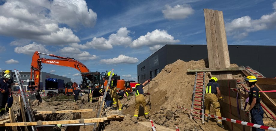 Feuerwehrleute in Schutzkleidung arbeiten gemeinsam an einer Übung im Bereich Technische Hilfeleistung am Bau. Im Hintergrund ein oragner Bagger.