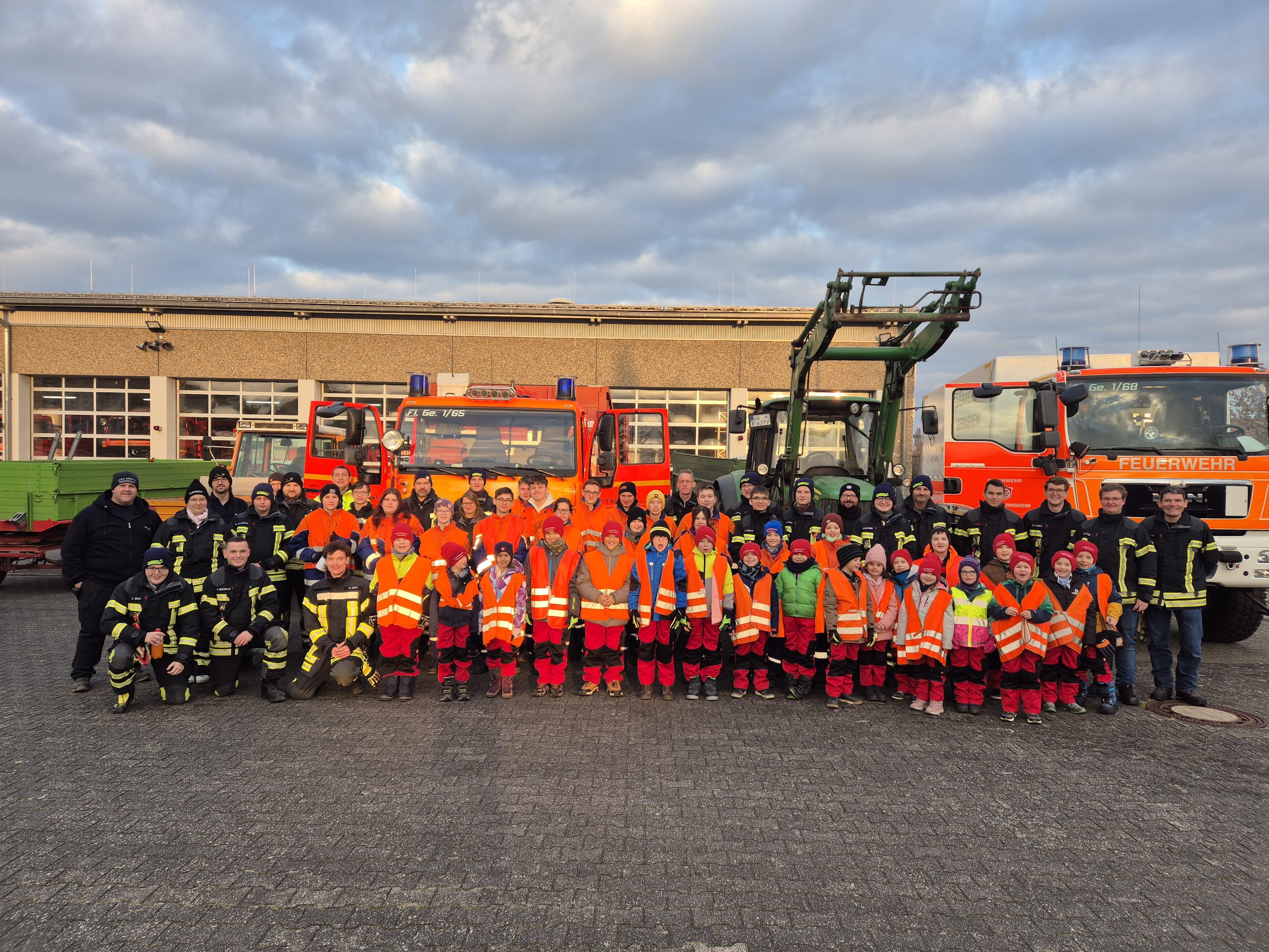 Große Gruppe von Feuerwehrleuten, darunter auch Kinder und Jugendliche, in Einsatz- und Warnkleidung vor mehreren Feuerwehrfahrzeugen vor einem Feuerwehrgerätehaus.
