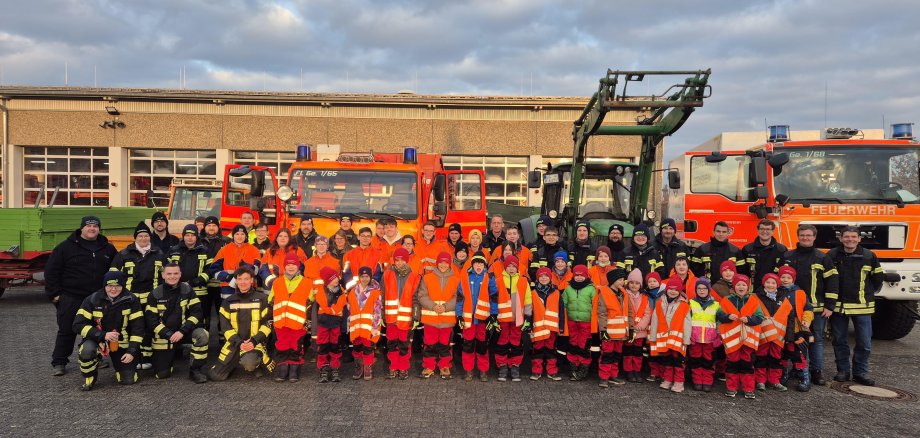 Große Gruppe von Feuerwehrleuten, darunter auch Kinder und Jugendliche, in Einsatz- und Warnkleidung vor mehreren Feuerwehrfahrzeugen vor einem Feuerwehrgerätehaus.