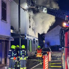 Feuerwehrleute stehen neben einer Drehleiter. Von der Drehleiter wird ein brennendes Haus gelöscht. Starke Rauchentwicklung aus dem Haus. Es ist Nacht.