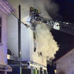 Feuerwehrleute stehen neben einer Drehleiter. Von der Drehleiter wird ein brennendes Haus gelöscht. Starke Rauchentwicklung aus dem Haus. Es ist Nacht.