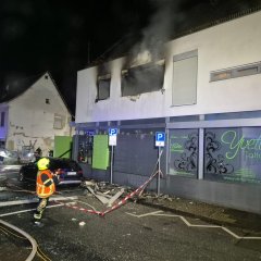 Ausgebranntes Wohnhaus bei Nacht. Im Vordergrund steht ein Feuerwehrmann und zwei Polizisten.