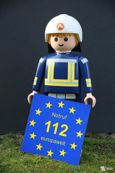 Ein Playmobil-Feuerwehrmann hält ein blaues Schild mit dem Text Notruf absetzen im Kreis darum sind Sterne.