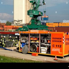 Ein geöffneter Abrollbehälter-Gefahrgut (AB-G). Daneben vier Feuerwehrleute. Im Hintergrund ein Containerhafen mit Kran.
