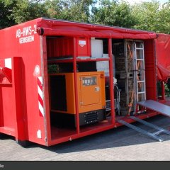 Roter Abrollbehälter der Feuerwehr für Hochwasserschutz (AB-HWS-2). Geöffnete Seite mit Blick auf eine Sandsackfüllmaschine.