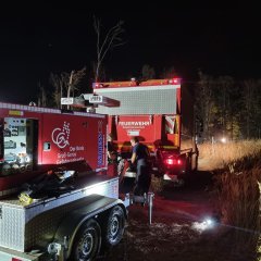 Roter Notstromanhänger hängt an einem Feuerwehrfahrzeug und steht auf einer Wiese. Im Hintergrund ein Waldstück.