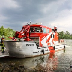Rotes Feuerwehrboot mit Namen Hecht, ein Hilfeleistungslöschboot (HLB), mit geöffneter Laderampe auf dem Rhein macht am Land fest. Im Hintergrund sind weitere kleine Boote der DLRG zu sehen.