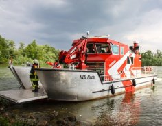Rotes Feuerwehrboot mit Namen Hecht, ein Hilfeleistungslöschboot (HLB), mit geöffneter Laderampe auf dem Rhein macht am Land fest. Im Hintergrund sind weitere kleine Boote der DLRG zu sehen.